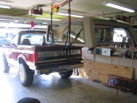 1996 Ford Bronco - A dark red full-size Ford SUV sits in a home garage with its tan fiberglass removable hardtop lifted off and suspended overhead on blue and…
