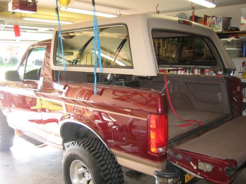 1996 Ford Bronco - Rear three-quarter view of a burgundy Ford Bronco in a garage with the tan hardtop suspended above the body on blue and red straps and the…