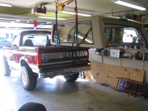 1996 Ford Bronco - A lifted burgundy Ford Bronco sits topless in a home garage while its tan fiberglass hardtop hangs above on blue ratchet straps and a ceili…