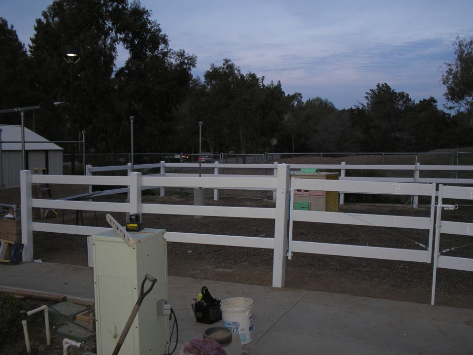 Outdoor scene with a white fence, a shed, a light pole, and various equipment including a shovel and a generator
