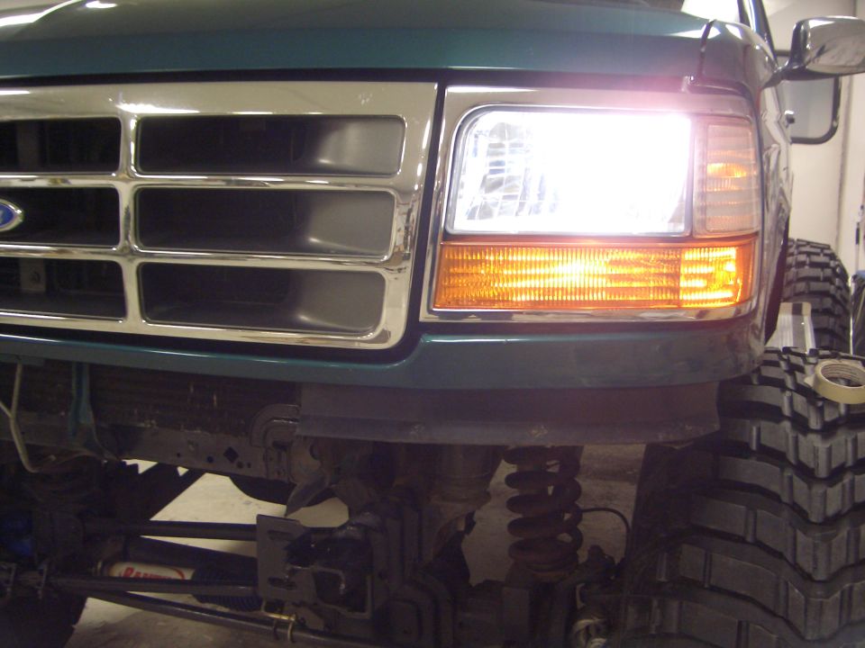 Front corner of green Ford Bronco with lit headlight, chrome grille, and lifted suspension with big tire