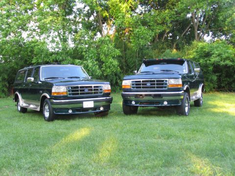 1995 Ford C-350 (Centurion Conversions) - Two dark green Ford Broncos are parked on a grassy lawn with trees behind them.