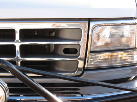 1994 Ford Bronco - Close-up of the front end of a chrome-trimmed grille and clear rectangular headlight assembly with amber turn signal on a light-colored tru…