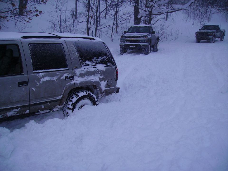 Silver SUV stuck in deep snow, with two pickup trucks behind it on a snowy road