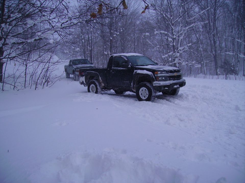 Black Chevrolet Colorado pickup truck stuck in snow, another truck behind it on a snowy road