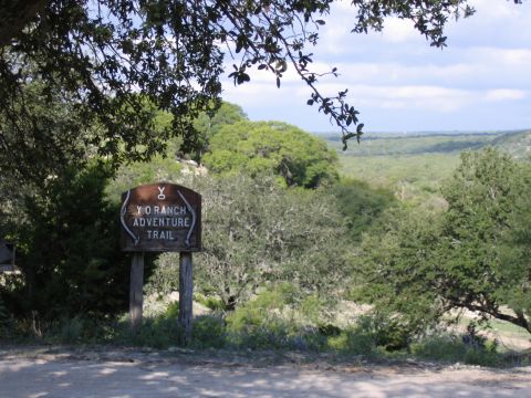 1996 Ford Bronco - A wooden sign marks the Y.O. Ranch Adventure Trail, set against a backdrop of trees and rolling hills under a partly cloudy sky.