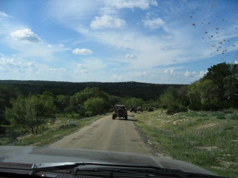 1996 Ford Bronco - A convoy of off-road vehicles travels down a rural road surrounded by trees and hills.