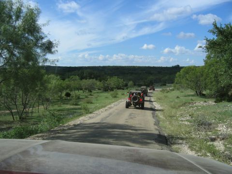 1996 Ford Bronco - A convoy of Jeeps travels down a rural road surrounded by green trees and open fields.