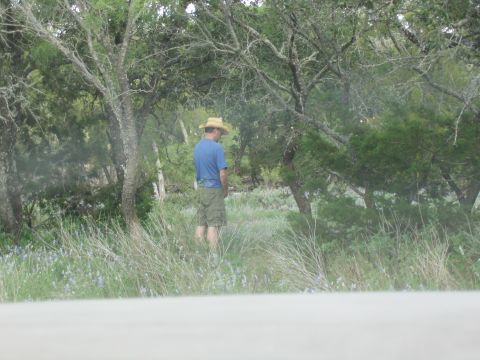 1996 Ford Bronco - A man wearing a straw hat and shorts is seen urinating in a wooded area with tall grass and small blue flowers.