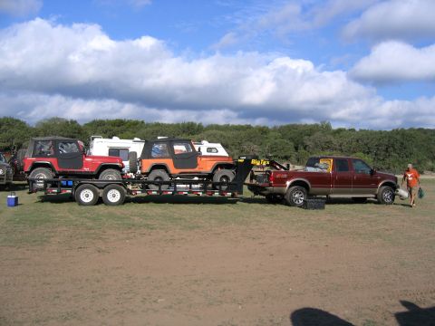 1996 Ford Bronco - A pickup truck tows a trailer carrying two Jeeps and a camper, with a person walking nearby.
