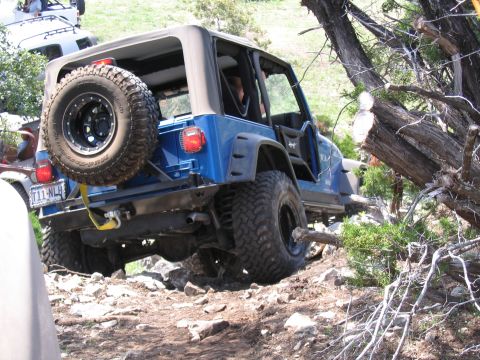 1996 Ford Bronco - Blue Jeep Wrangler navigating a challenging rocky trail with a spare tire mounted on the rear.