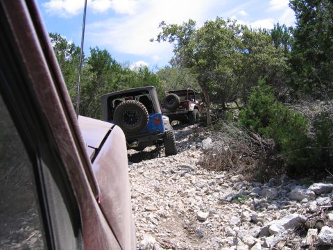 1996 Ford Bronco - Several Jeeps navigate a challenging, off-road trail with a rocky surface and dense vegetation.