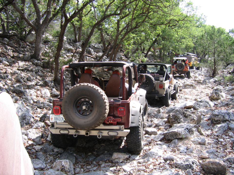 Several Jeeps driving single file on a rough, rocky path through a wooded area. The lead Jeep is dark red, followed by a tan one, then an orange one