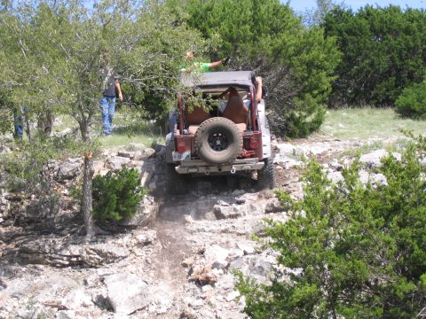 1996 Ford Bronco - A red Jeep Wrangler navigates a steep, rocky incline surrounded by trees and brush.