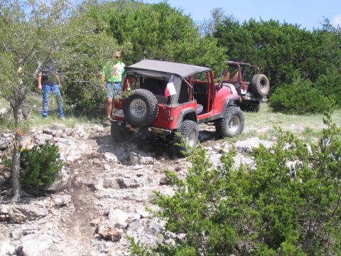 1996 Ford Bronco - Two red Jeep Wranglers navigate a rocky off-road trail surrounded by trees and brush.