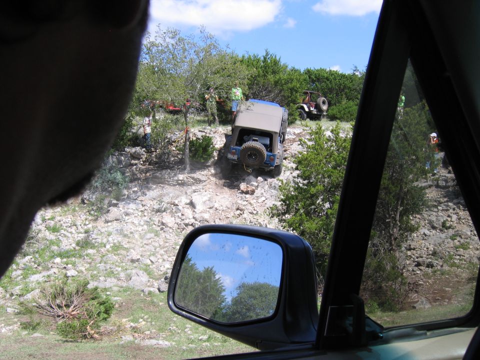 Blue Jeep Wrangler on rocky incline with spare tire on rear door, other off-road vehicles visible