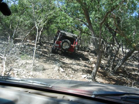 1996 Ford Bronco - A red Jeep navigates a challenging off-road trail surrounded by trees, with another vehicle partially visible further up the path.