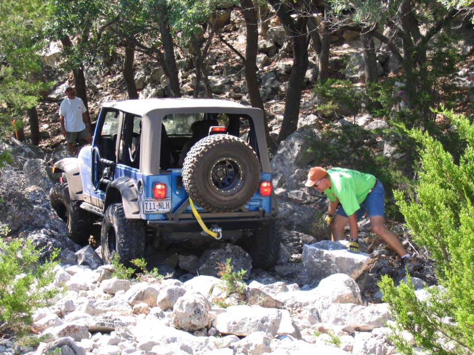 Blue Jeep Wrangler with spare tire on rear, navigating a rocky trail