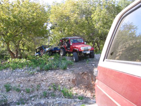 1996 Ford Bronco - Two Jeeps are seen off-road, with one overturned, surrounded by trees and rocky terrain.