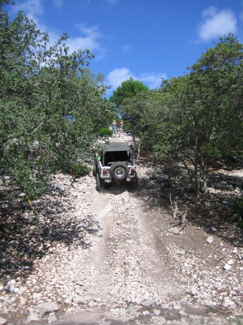 1996 Ford Bronco - A silver Jeep Wrangler navigates a steep, rocky trail with other vehicles and people ahead on the path.