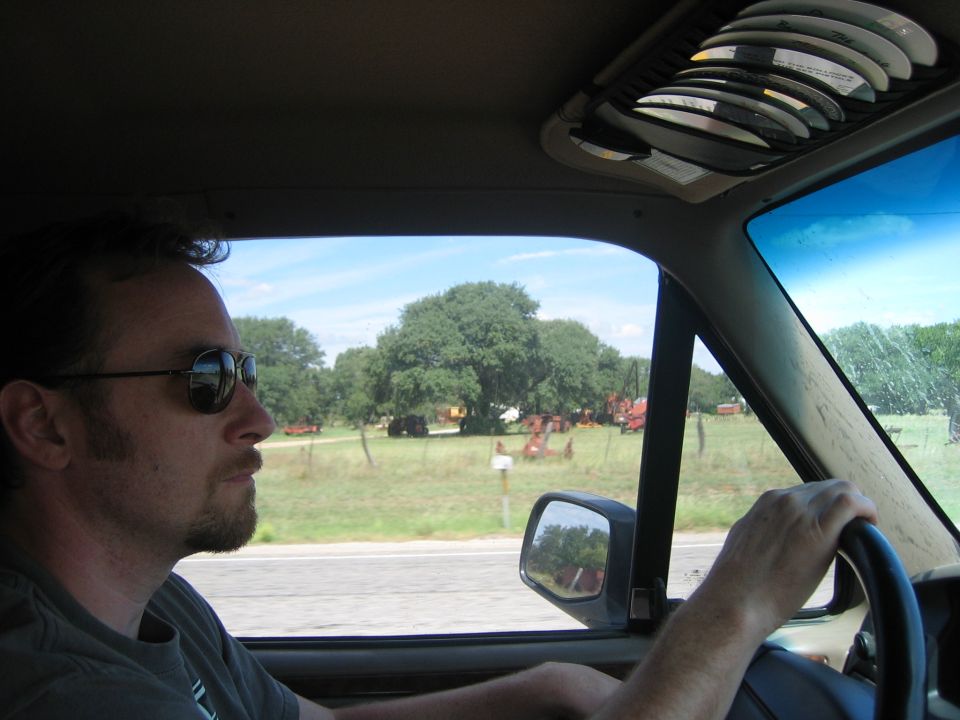 Interior view of a vehicle with a man driving, looking out at farm equipment in a field
