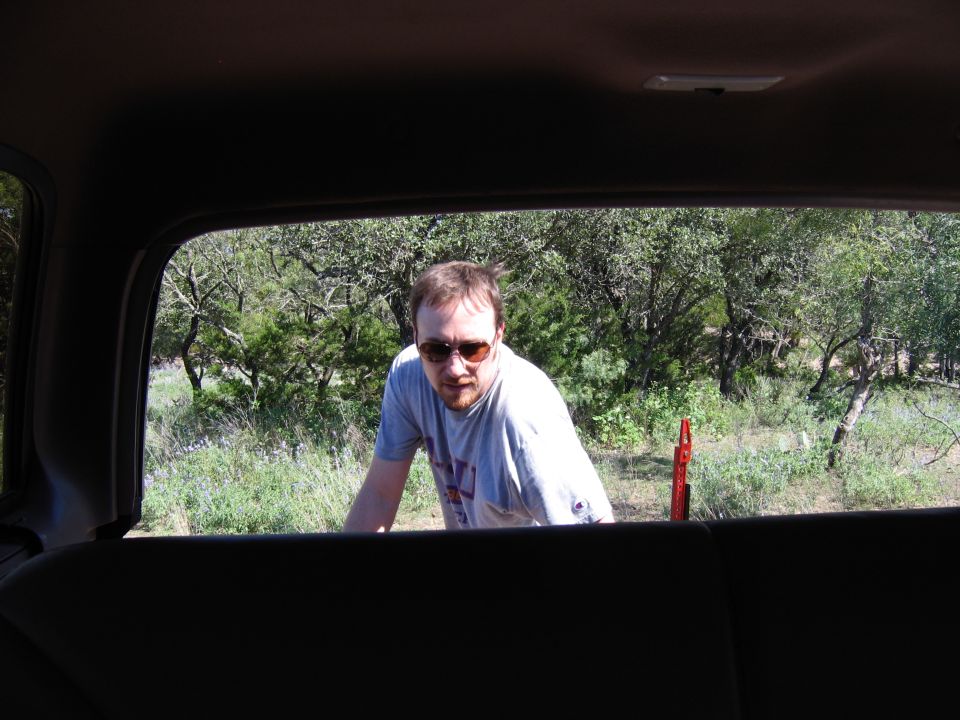 Man with sunglasses leans out of a dark interior, looking towards a wooded area with green foliage and a red post