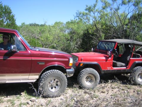 1996 Ford Bronco - A red 1996 Ford Bronco is seen parked next to a red Jeep Wrangler on a dirt road with trees in the background.