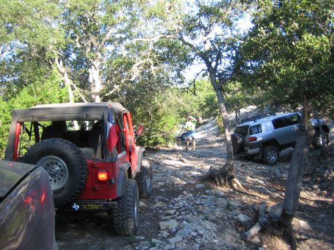1996 Ford Bronco - Two off-road vehicles navigate a rocky trail surrounded by trees, with a red Jeep Wrangler in the foreground and a silver Toyota FJ Cruiser…