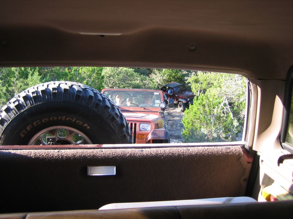 Spare tire in foreground, two Jeeps on a dirt road in background, surrounded by trees