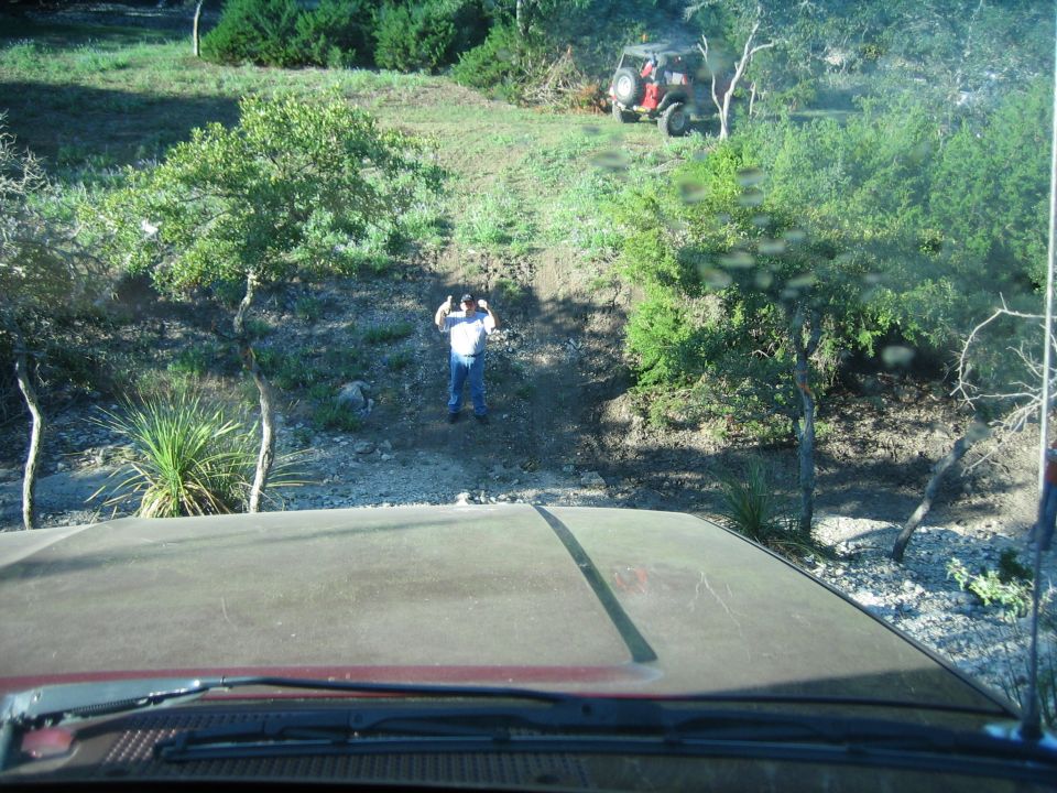 Red vehicle hood in foreground, person standing on dirt path, red Jeep on hill