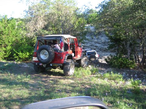 1996 Ford Bronco - Red Jeep Wrangler on a dirt path, with another vehicle partially visible behind it.