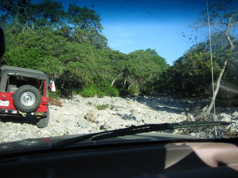 1996 Ford Bronco - A red Jeep Wrangler navigates a rocky trail surrounded by dense foliage under a clear blue sky.