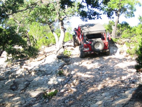 1996 Ford Bronco - A red Jeep navigates a challenging off-road trail with rocky terrain and sparse trees.