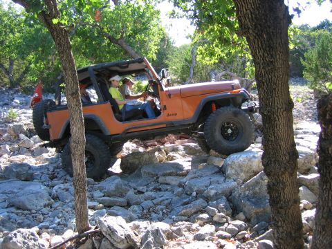 1996 Ford Bronco - An orange Jeep Wrangler is shown off-roading on a rocky terrain with trees surrounding the area.