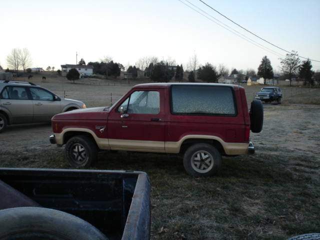 Red 1986 Ford Bronco II parked outside, spare tire mounted on rear door. Another SUV is parked to its left