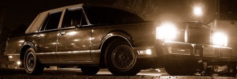 1989 Lincoln Town Car - A dark 1989 Lincoln Town Car is illuminated by headlights at night, viewed from a low 3/4 front angle.
