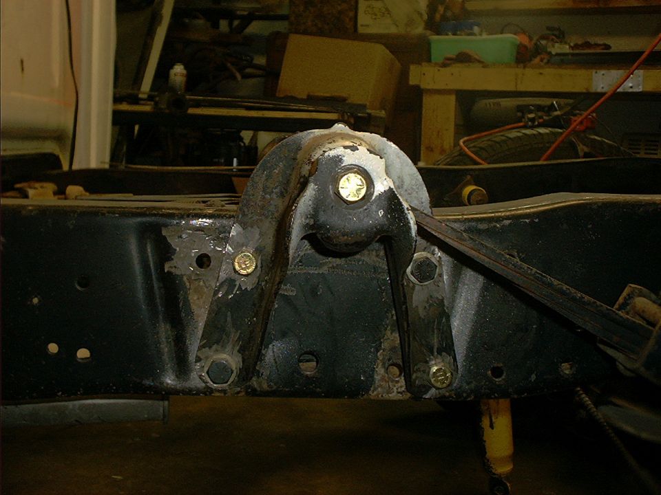 Close-up of black frame spring hanger with new gold bolts and leaf spring on a bare truck chassis in a garage