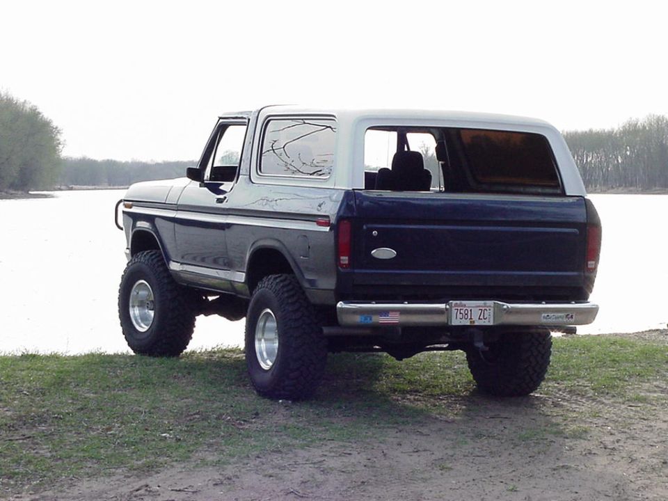 Two-tone blue and silver lifted Ford Bronco, rear 3/4 view by a lake