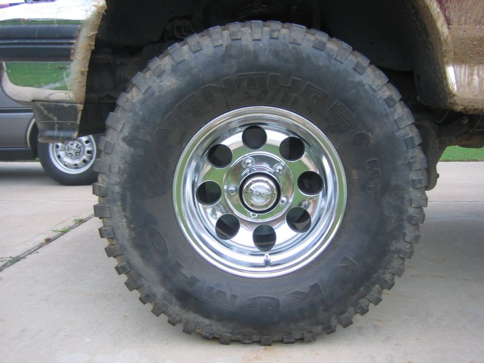 Close-up of a large off-road tire on a chrome wheel. Another vehicle is visible in the background