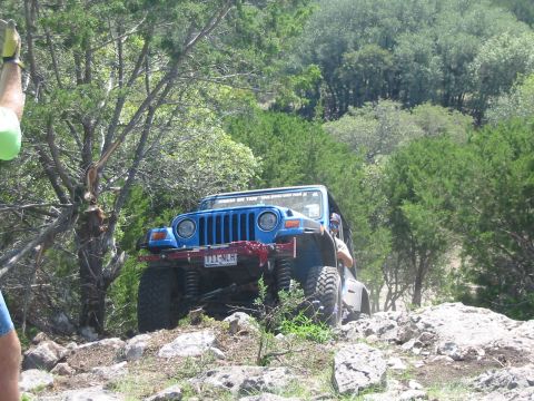 1996 Ford Bronco - A blue Jeep Wrangler navigates a rocky off-road trail surrounded by trees.