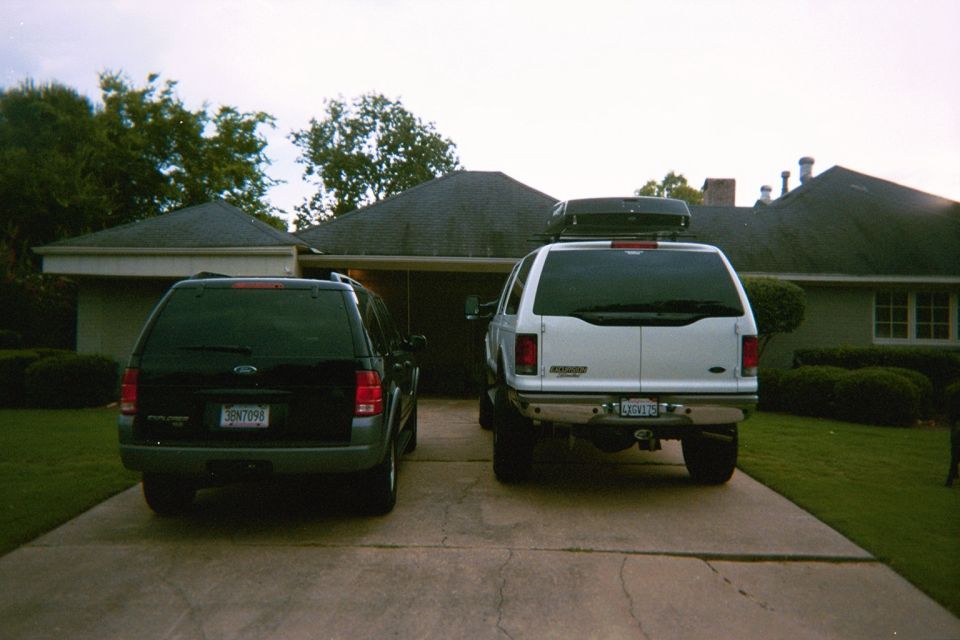 White lifted Ford Excursion with roof box parked beside a green Ford Explorer in a driveway