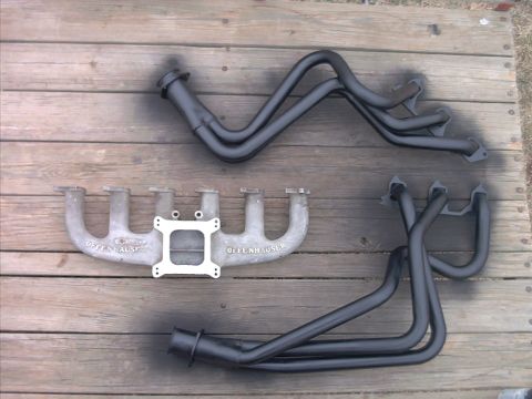 1985 Ford Bronco - Two exhaust manifolds, one aluminum and one cast iron, and a set of black headers are laid out on a wooden deck.