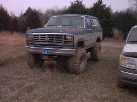 1985 Ford Bronco - A lifted Ford Bronco sits in a field next to another vehicle under an overcast sky.