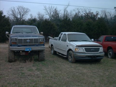 1985 Ford Bronco - A lifted blue Ford F-Series truck is parked in a grassy field next to a white pickup.