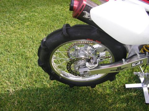 1900 Honda Big Red - Close-up of a motorcycle's rear wheel and exhaust system, set against a grassy background.