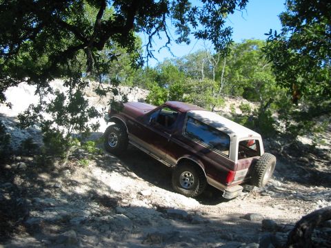 1996 Ford Bronco - A maroon 1996 Ford Bronco navigates a challenging, rocky off-road path under a canopy of trees.
