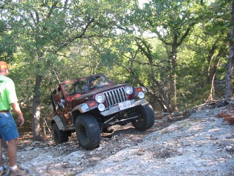 1996 Ford Bronco - A red Jeep Wrangler is shown off-roading on a rocky path surrounded by trees, with a person observing from the side.