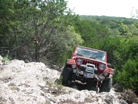 1996 Ford Bronco - A red Jeep Wrangler navigates a rocky off-road trail surrounded by trees and brush.