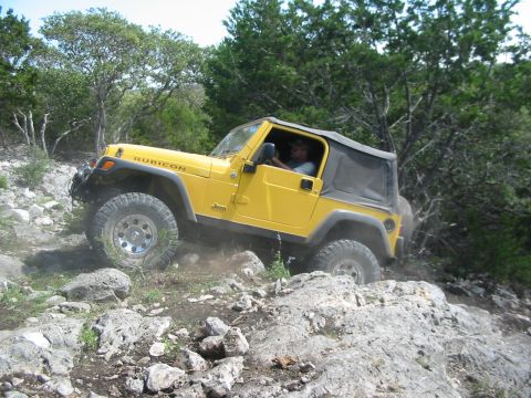 1996 Ford Bronco - Yellow Jeep Wrangler Rubicon navigating a rocky trail with trees in the background.