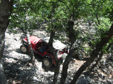 1996 Ford Bronco - A red off-road vehicle is seen traversing a challenging, rocky path surrounded by trees.